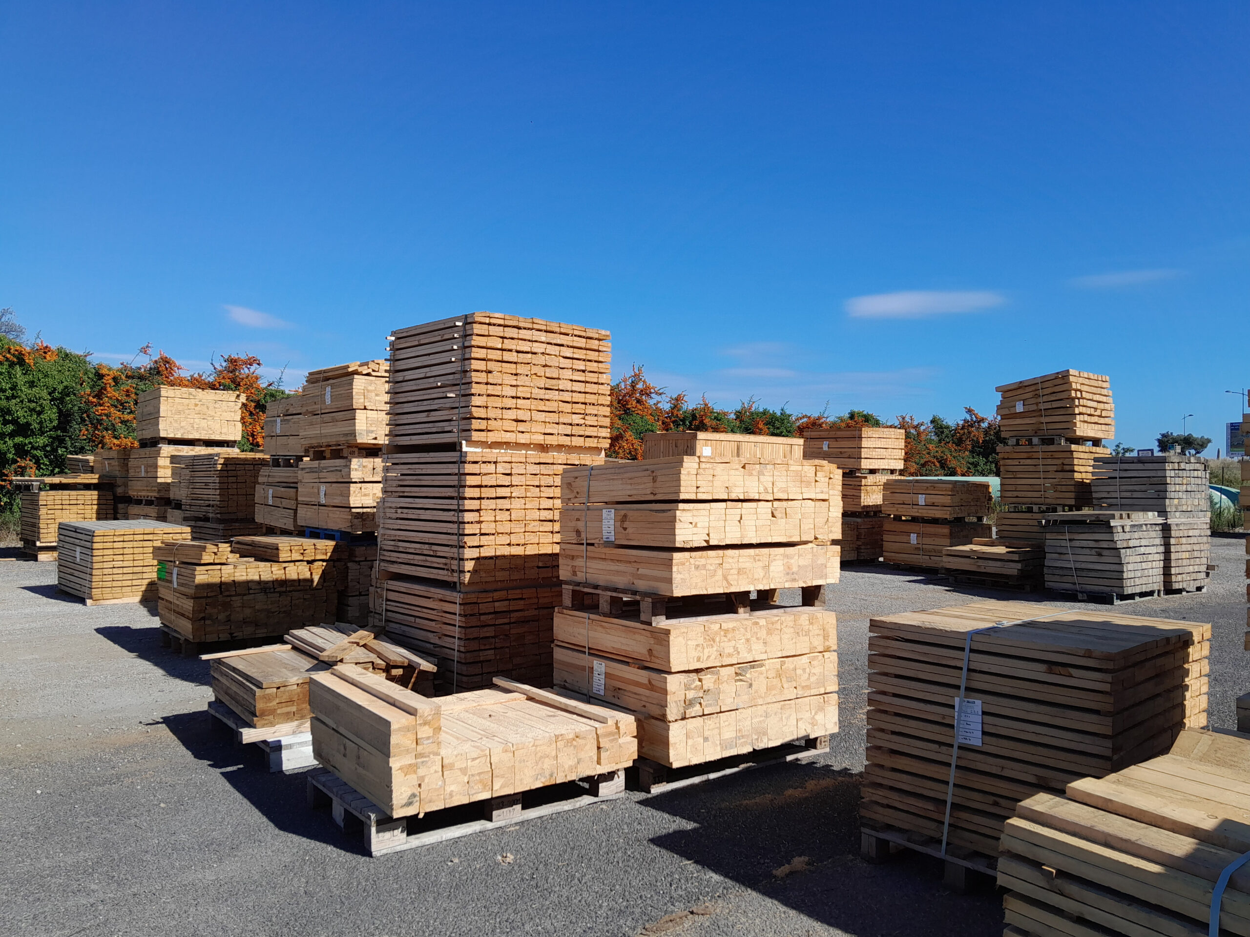 Grandes piles de planches de bois sciées et de palettes, stockées en extérieur sous un ciel bleu.